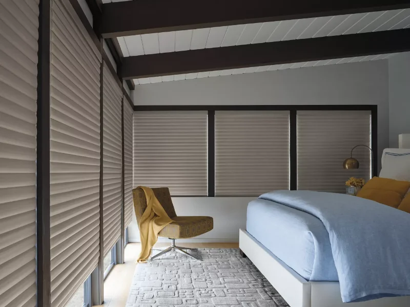 Beige cellular shades covering the large windows of a bedroom.